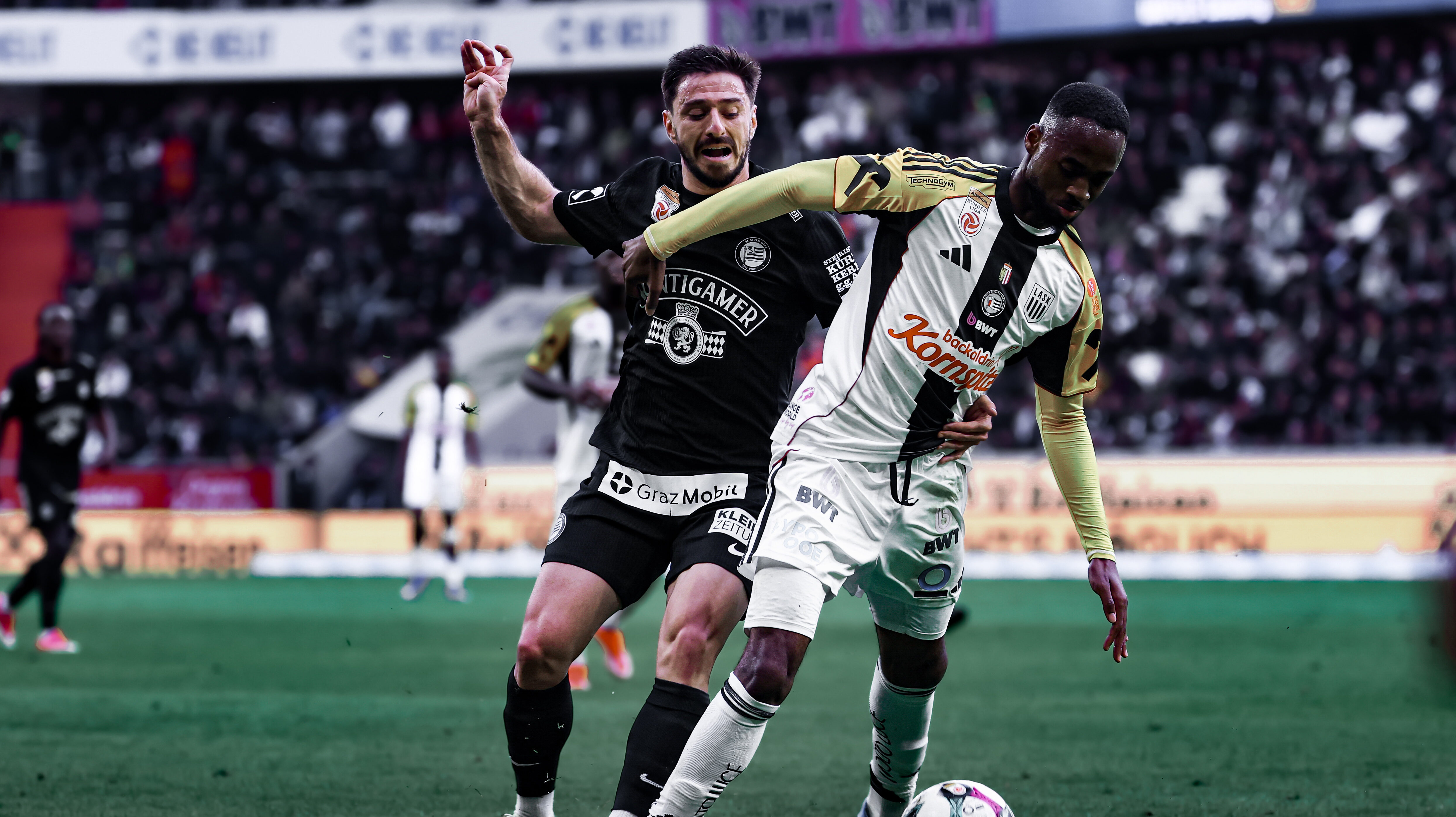 Ein Spieler in schwarz und einer in weiß-schwarz-gelb kämpfen um einen weiß-schwarzen Ball.