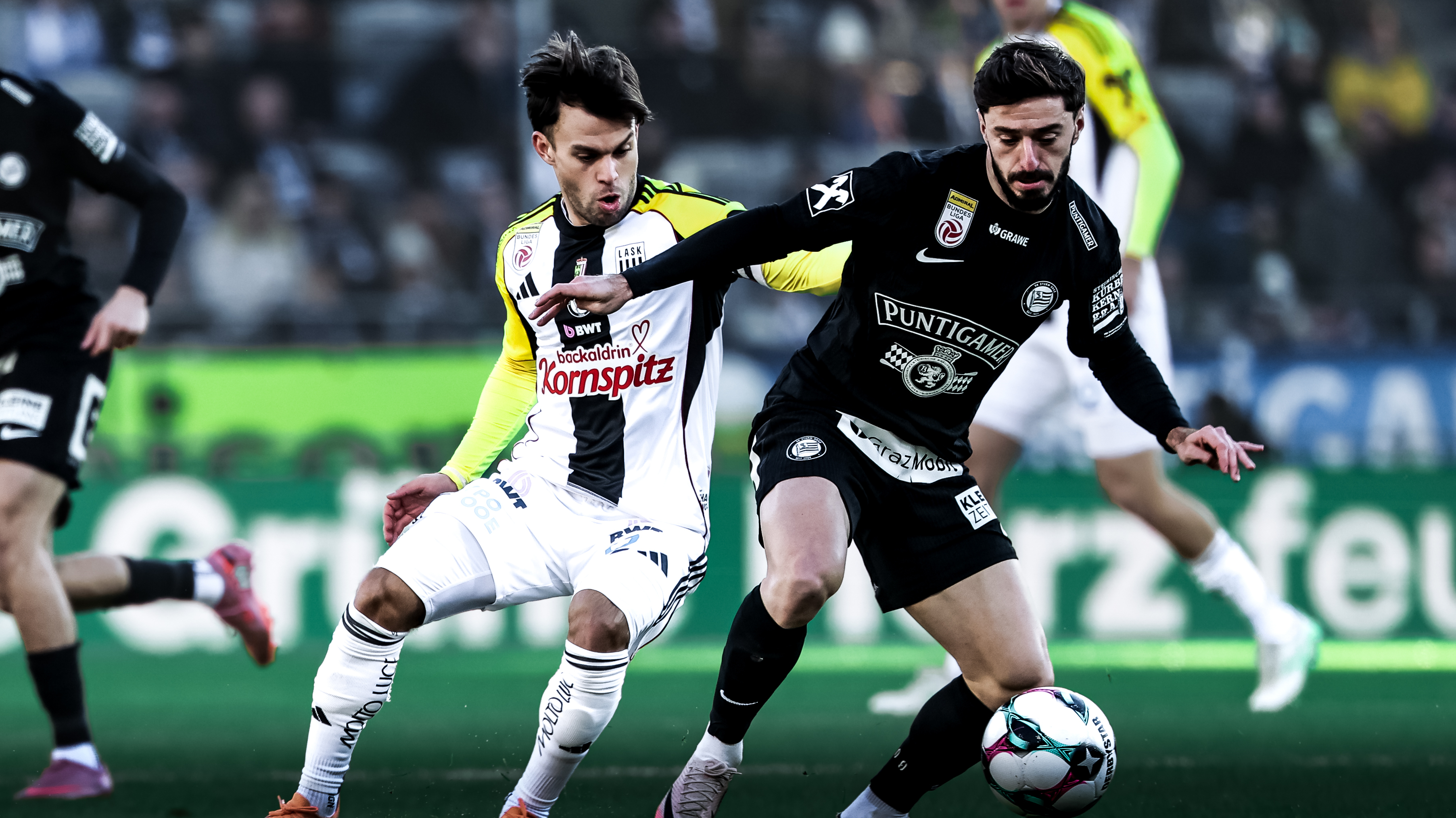 Ein Spieler in einem schwarzen Trikot kämpft mit einem Spieler in einem weiß-schwarz-gelben Trikot um einen weißen Ball