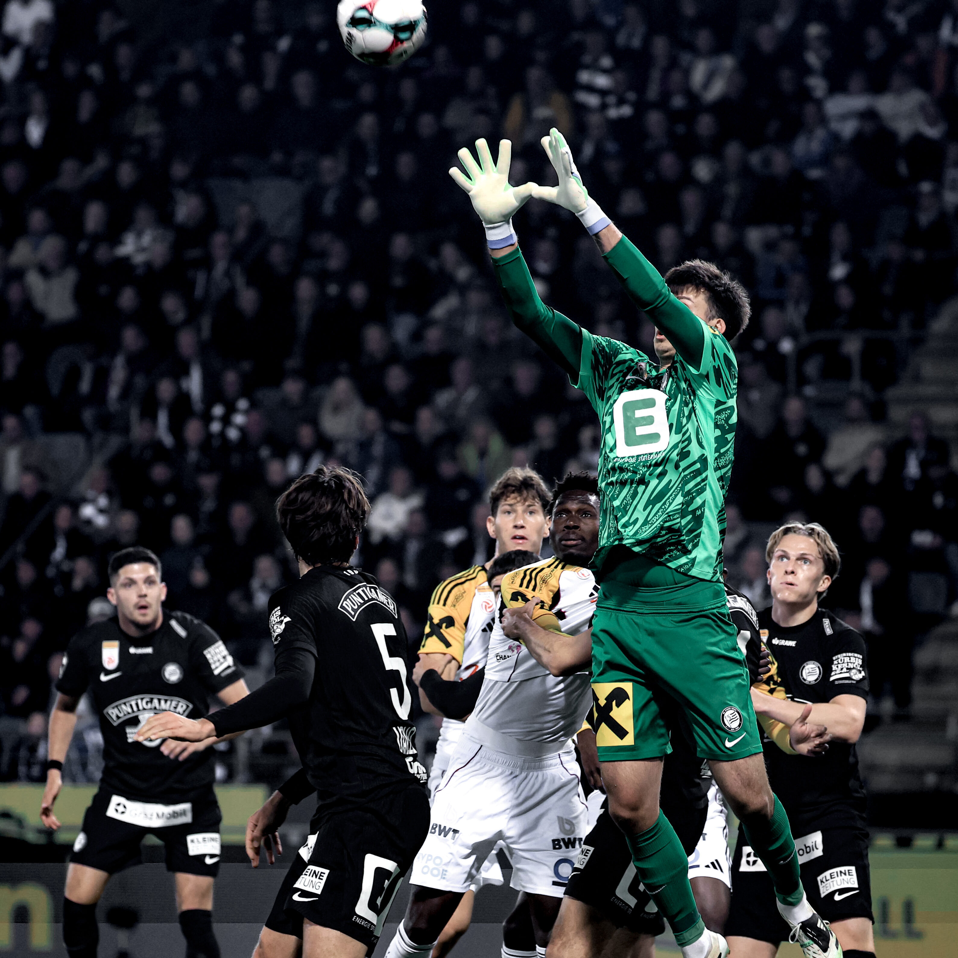 Ein Torhüter in grün fängt umringt von Spielerin in weiß und schwarz einen weißen Ball herunter.