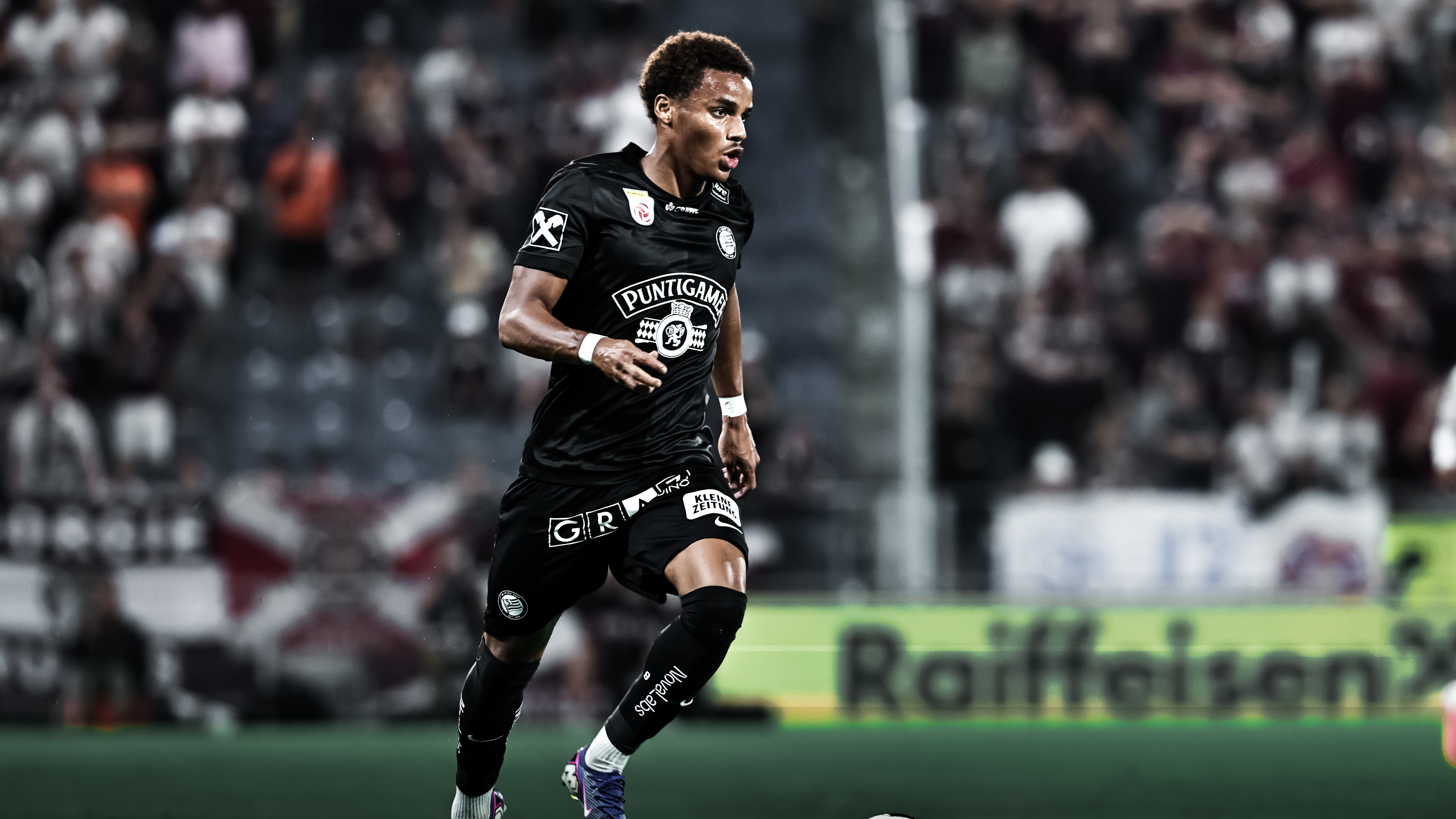 Ein Spieler in einem schwarzen Trikot läuft und schaut nach rechts, im Hintergrund sind Fans zu sehen.