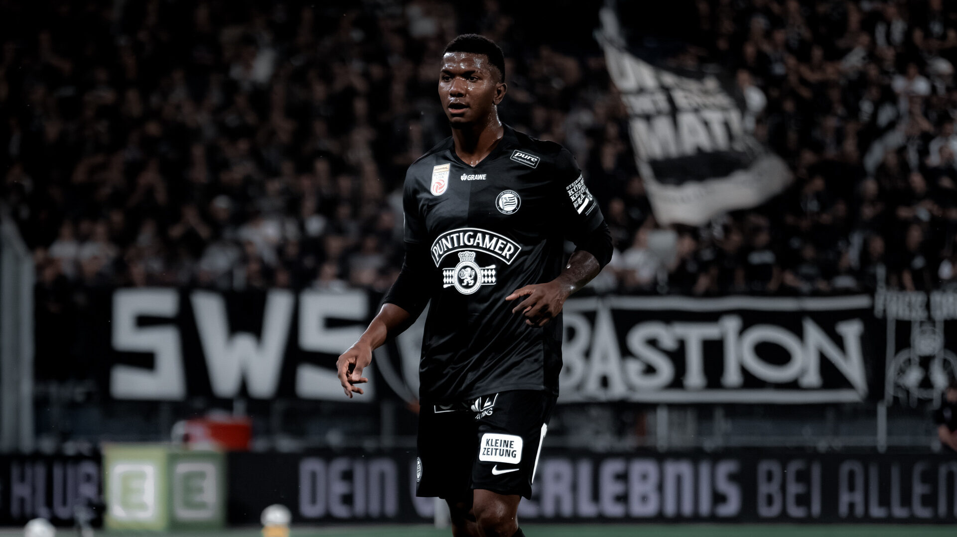 Ein Spieler in einem schwarzen Shirt steht auf einem grünen Rasen und schaut in die Ferne. Im Hintergrund sind Fans zu sehen.