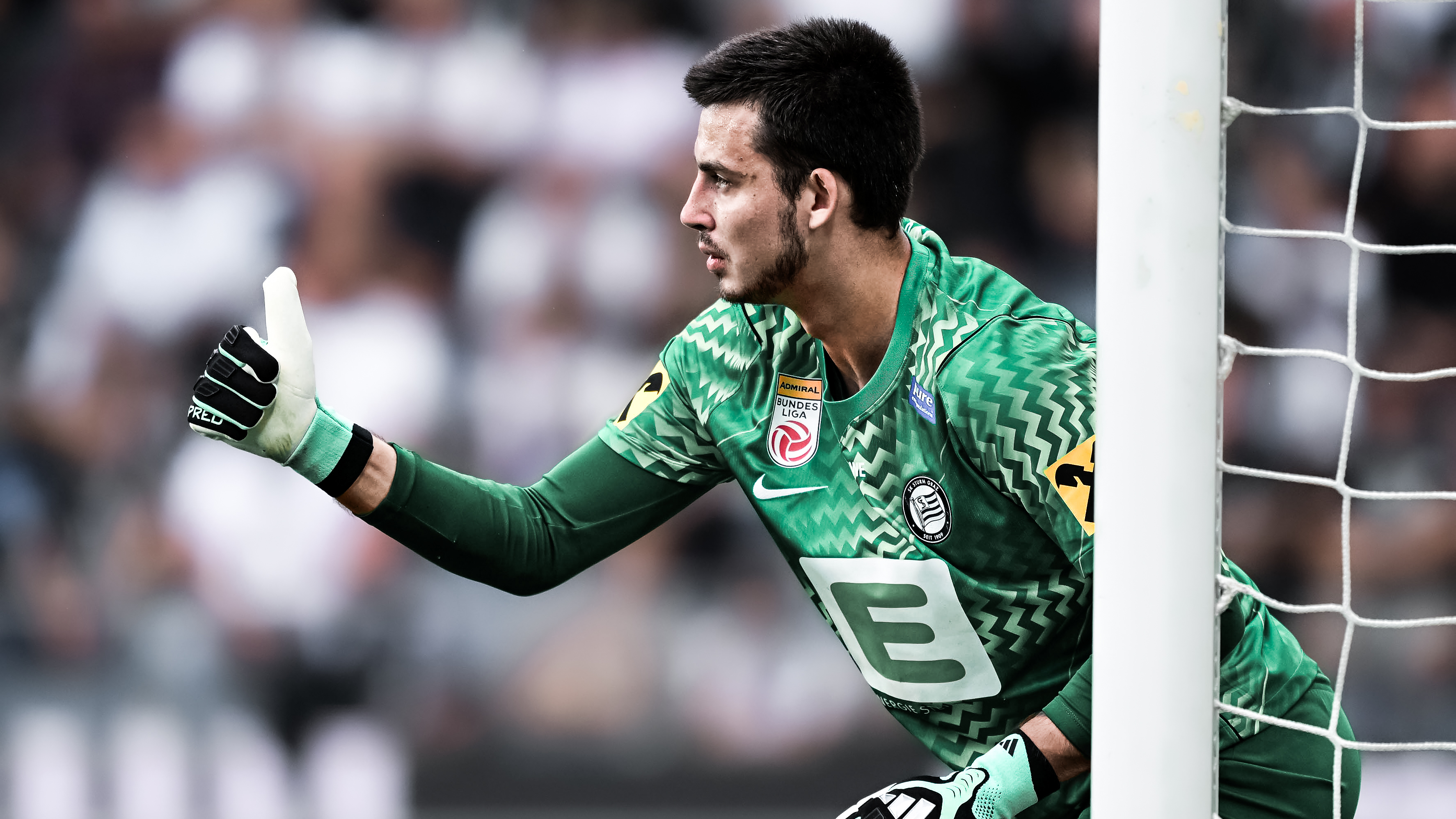 Ein Spieler in einem grünen Trikot mit Tormannhandschuhen an den Händen zeigt mit dem Daumen nach oben. Rechts im Bild ist eine Torstange und ein Netz zu sehen.