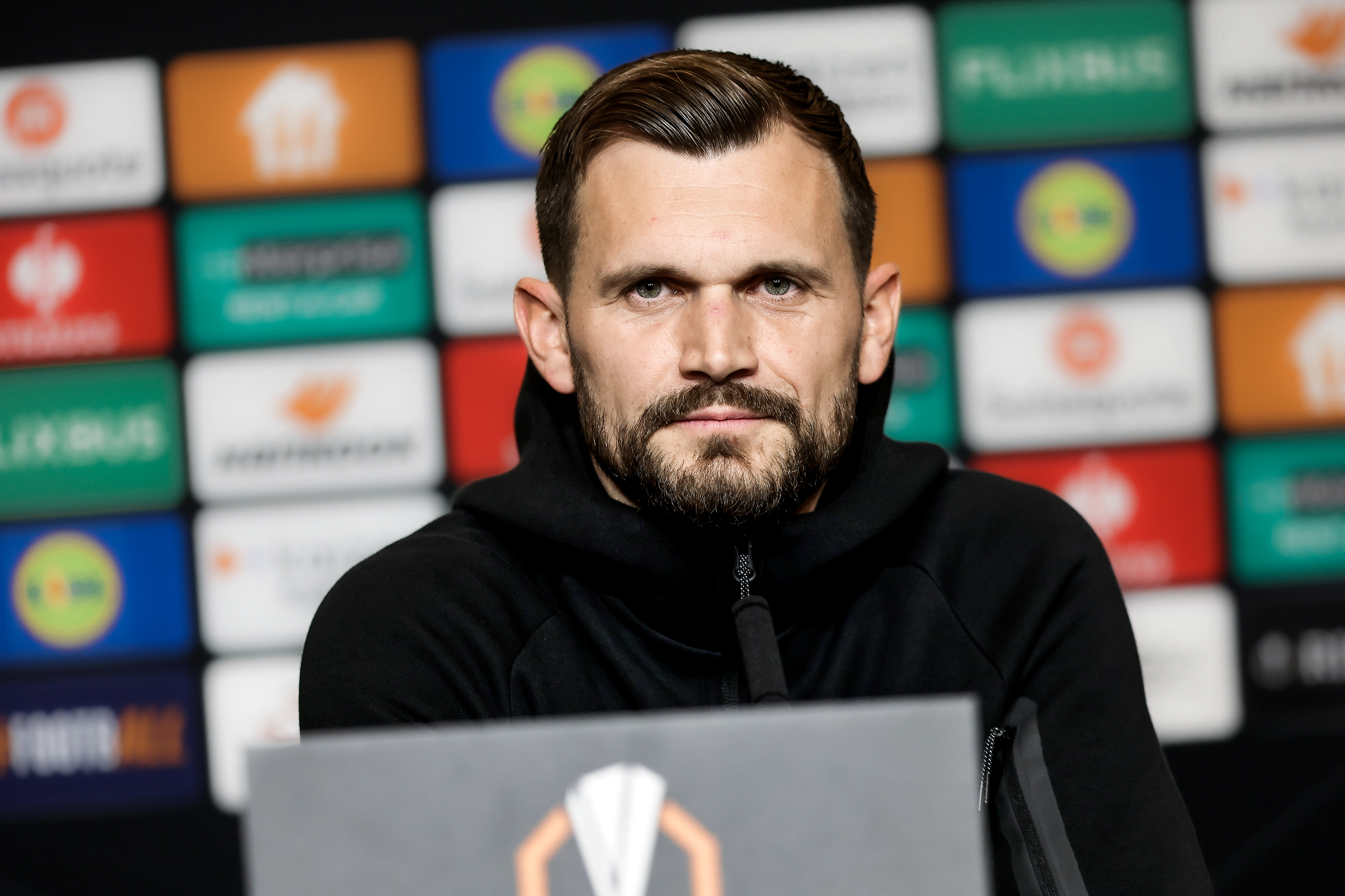 Ein Mann in schwarzer Weste sitzt vor einem Mikro und schaut.