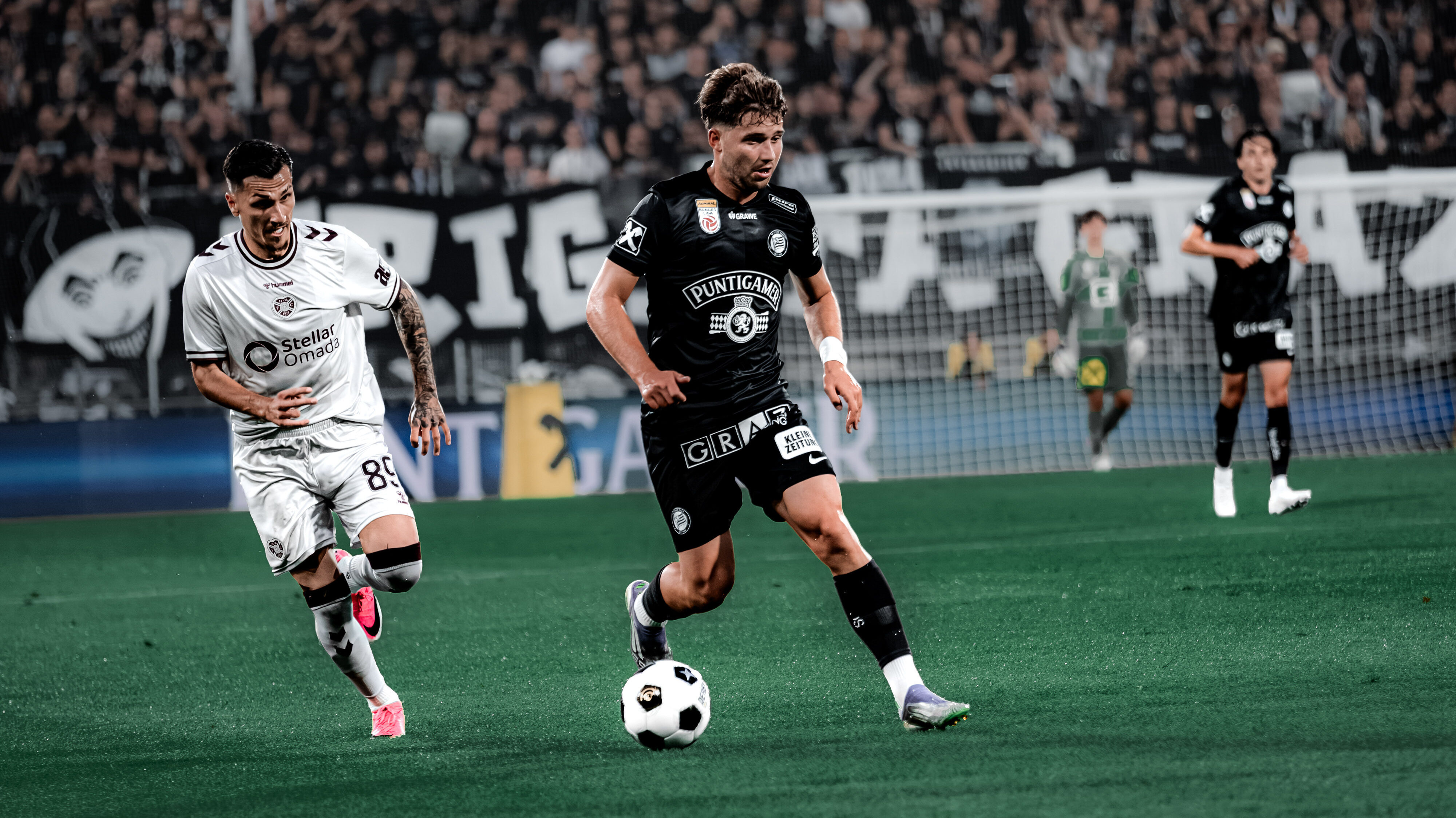 Ein Spieler in schwarz kämpft mit einem Spieler in weiß um einen weißen Ball. Im Hintergrund sind viele Menschen auf einer Tribüne zu sehen.