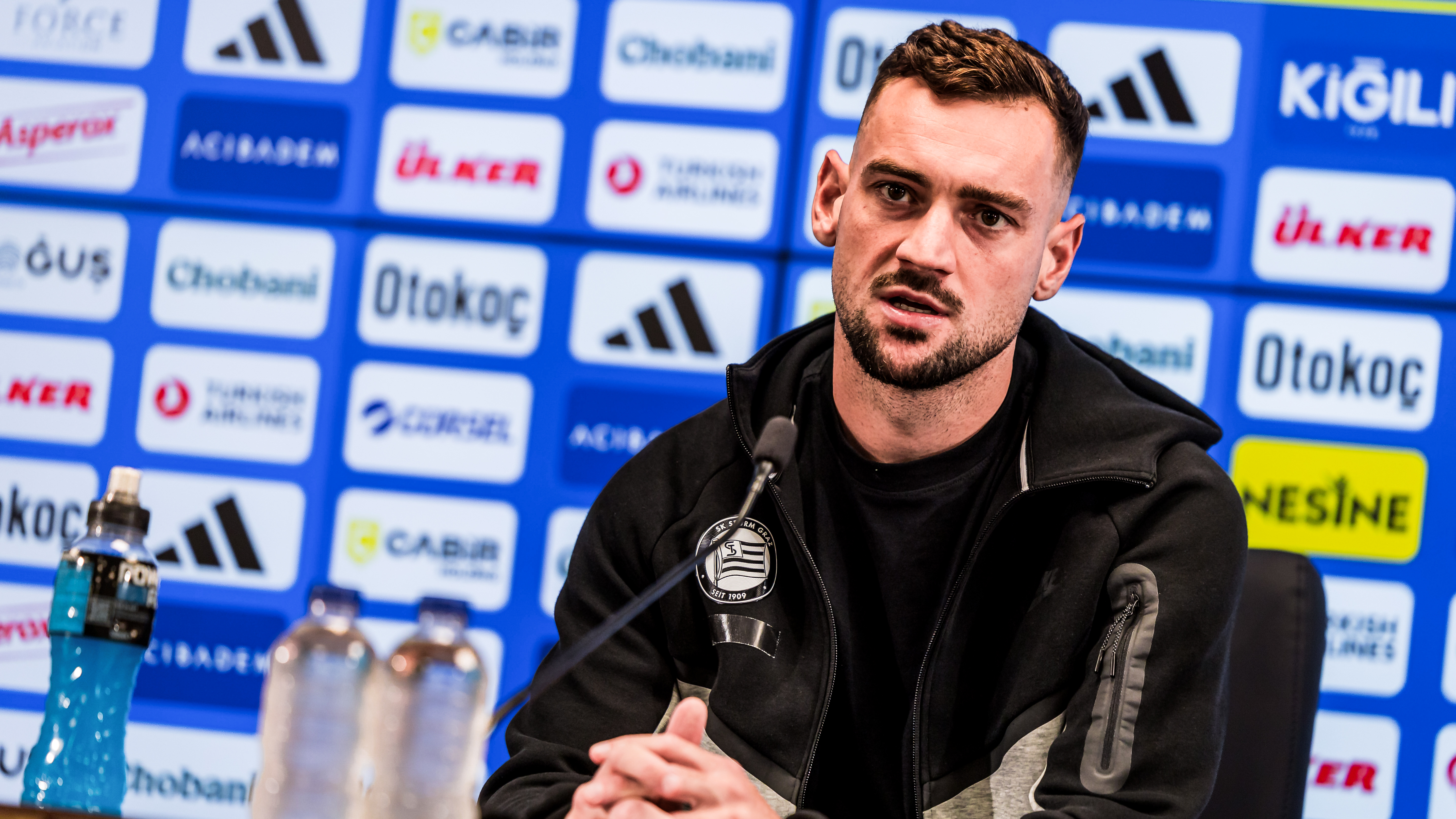 Ein Mann in schwarzen Gewand sitzt vor einer blauen Wand voller Logos