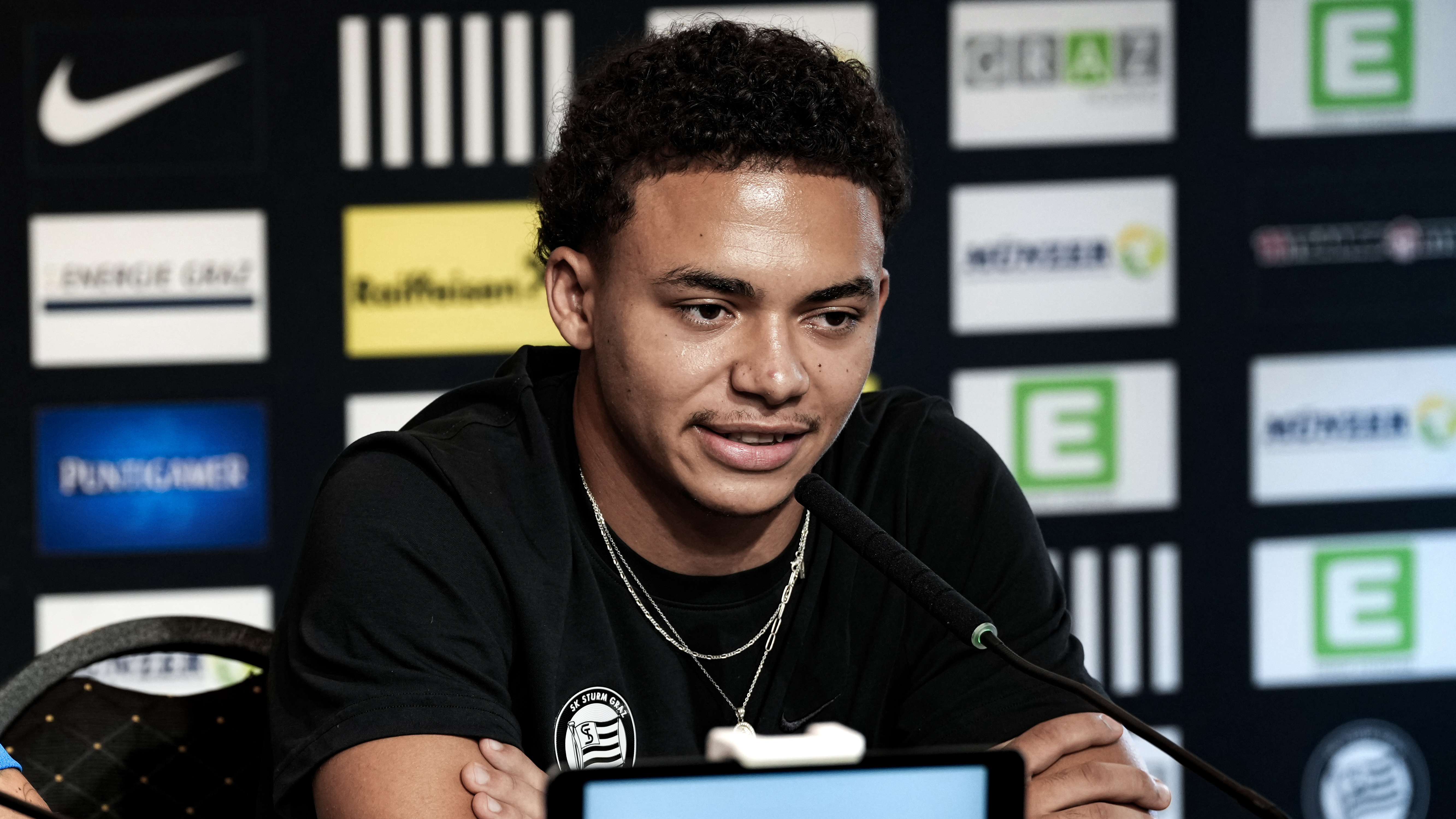 Ein Spieler sitzt in einem schwarzen Shirt vor einer schwarzen Sponsorenwand und spricht in ein Mikrofon