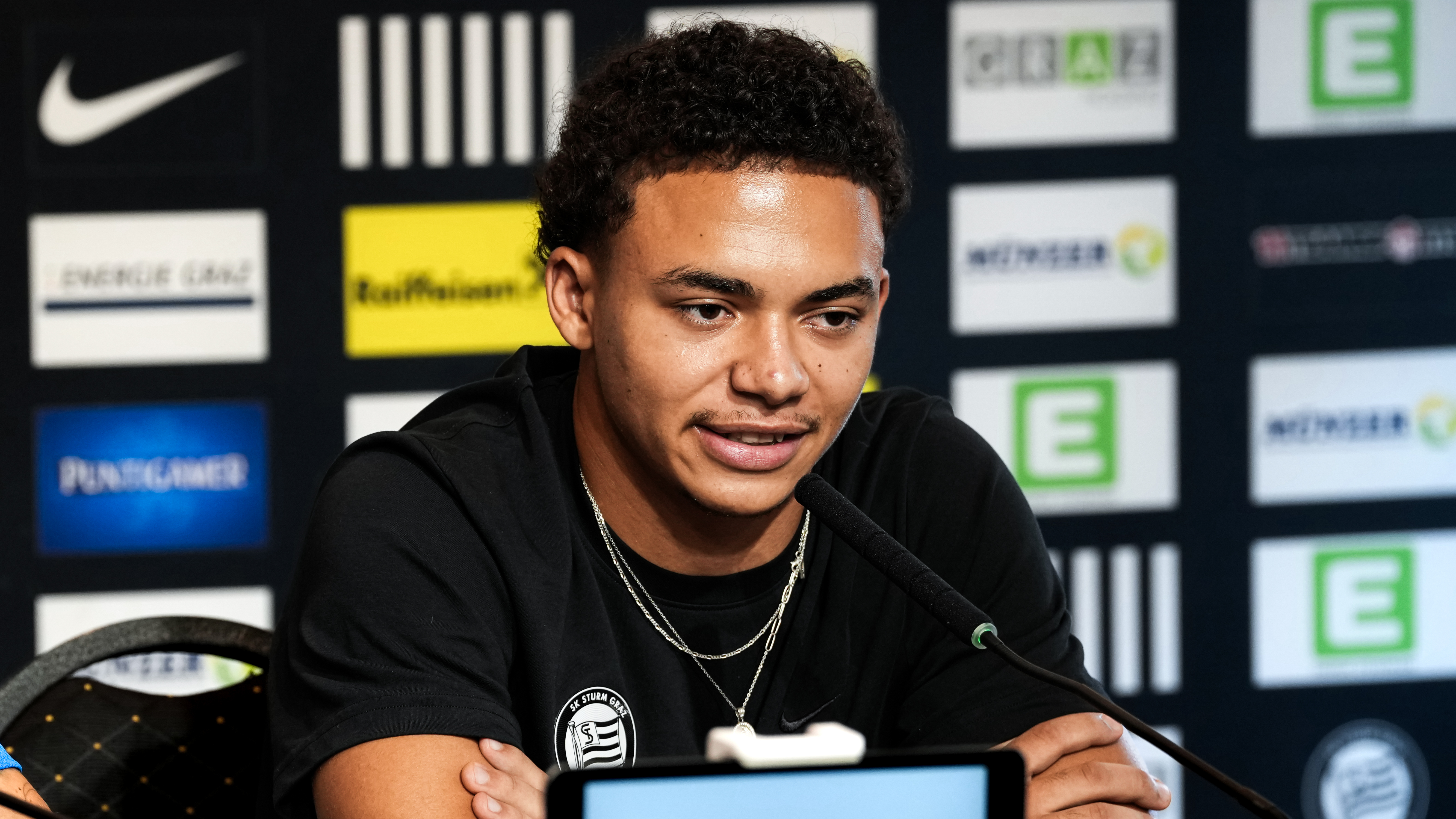 Ein Spieler sitzt in einem schwarzen Shirt vor einer schwarzen Sponsorenwand und spricht in ein Mikrofon