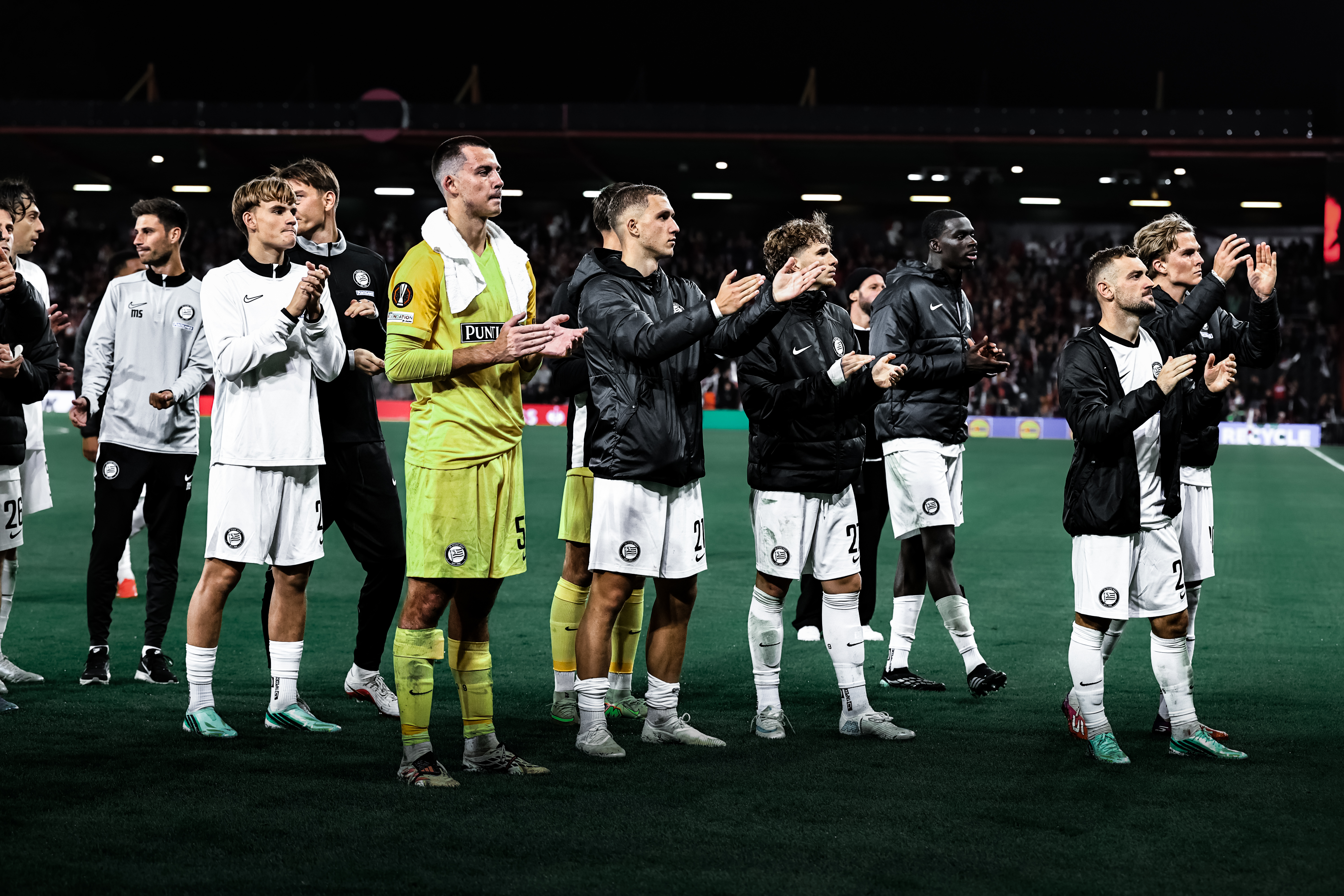 Zehn Spieler mit schwarzen Jacken und weißen Stutzen stehen auf einem grünen Rasen und klatschen