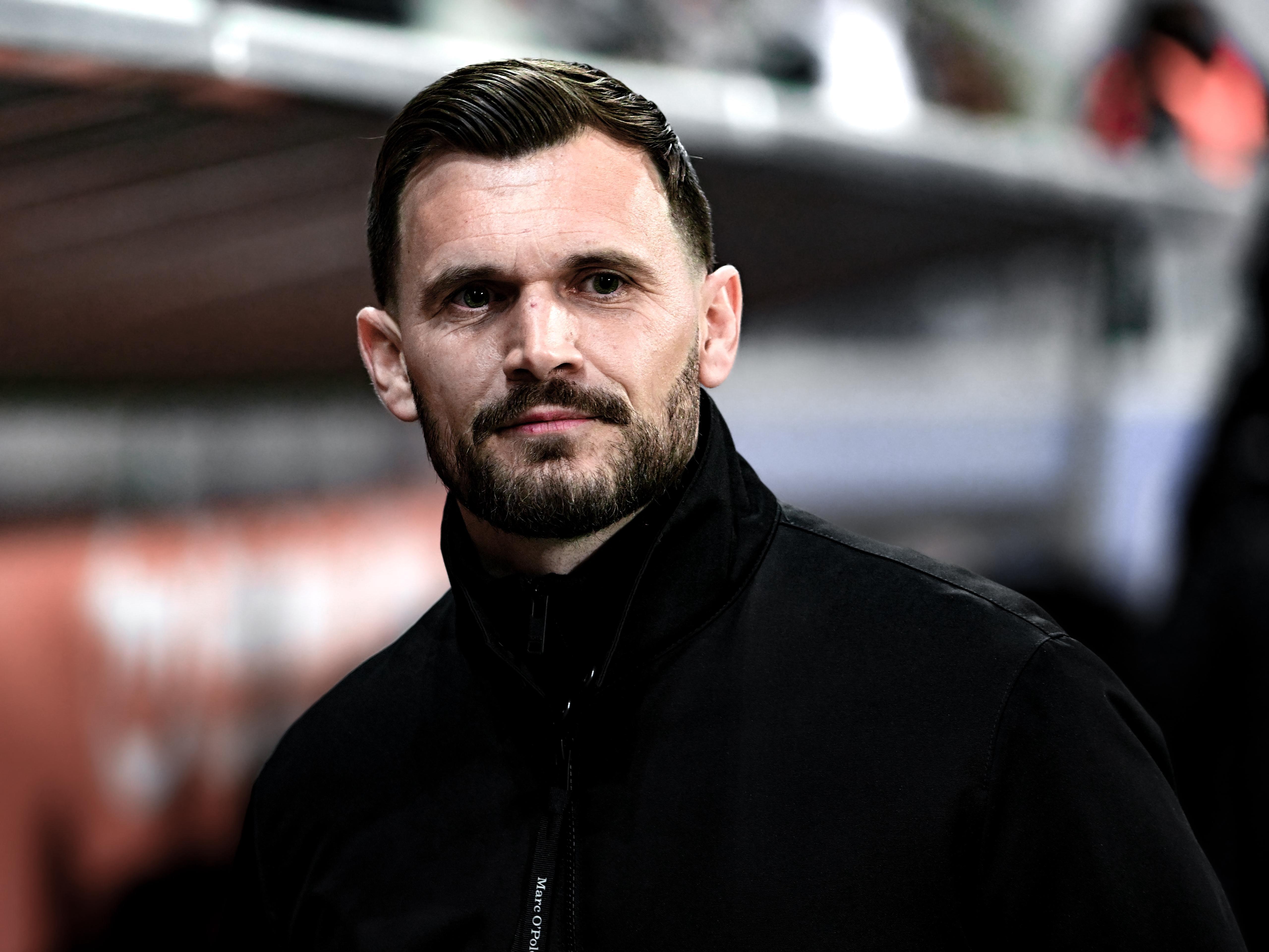 Mann mit dunkler Jacke steht vor einer Trainerbank.