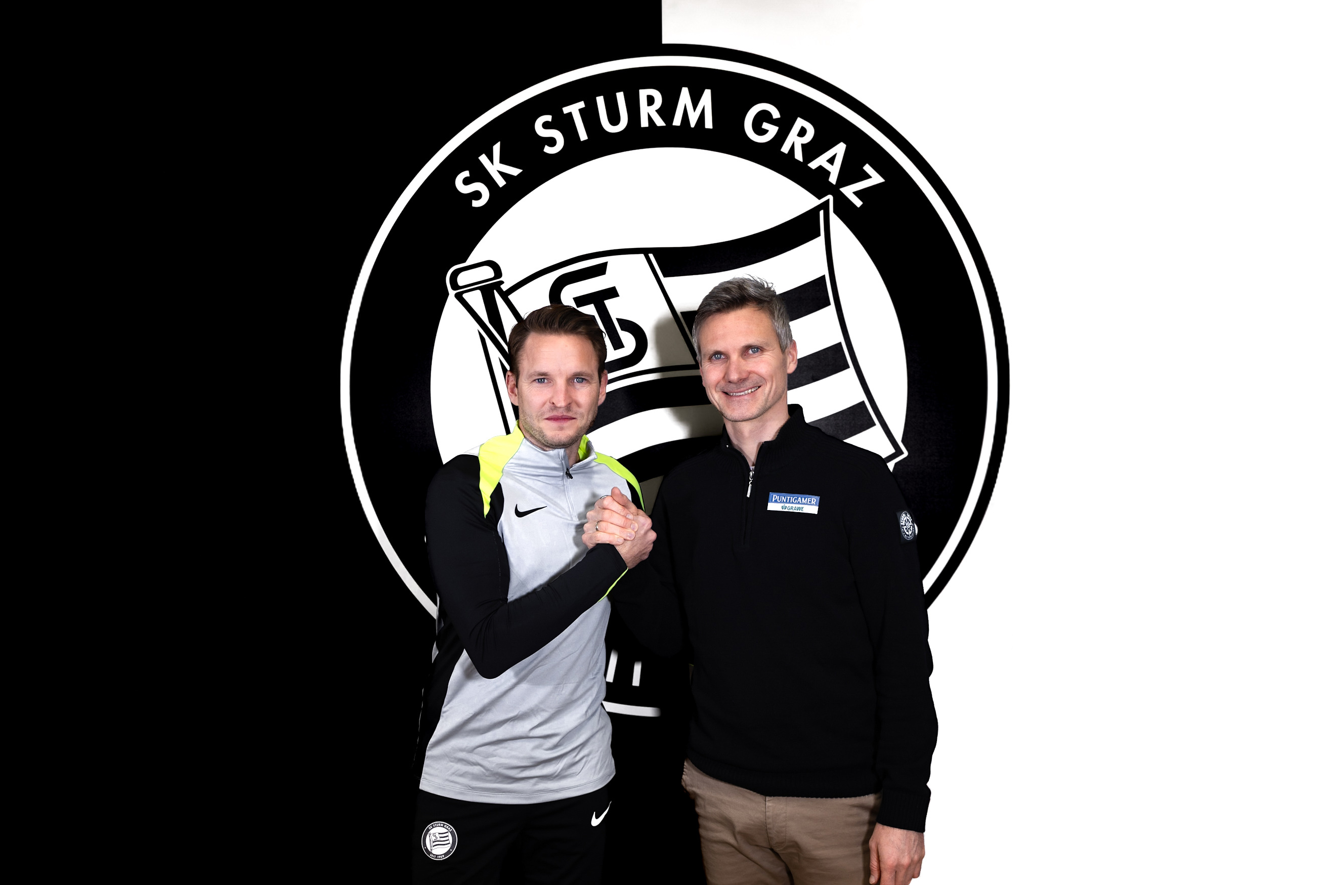 Zwei Personen stehen vor einem Logo und geben sich einen Handschlag