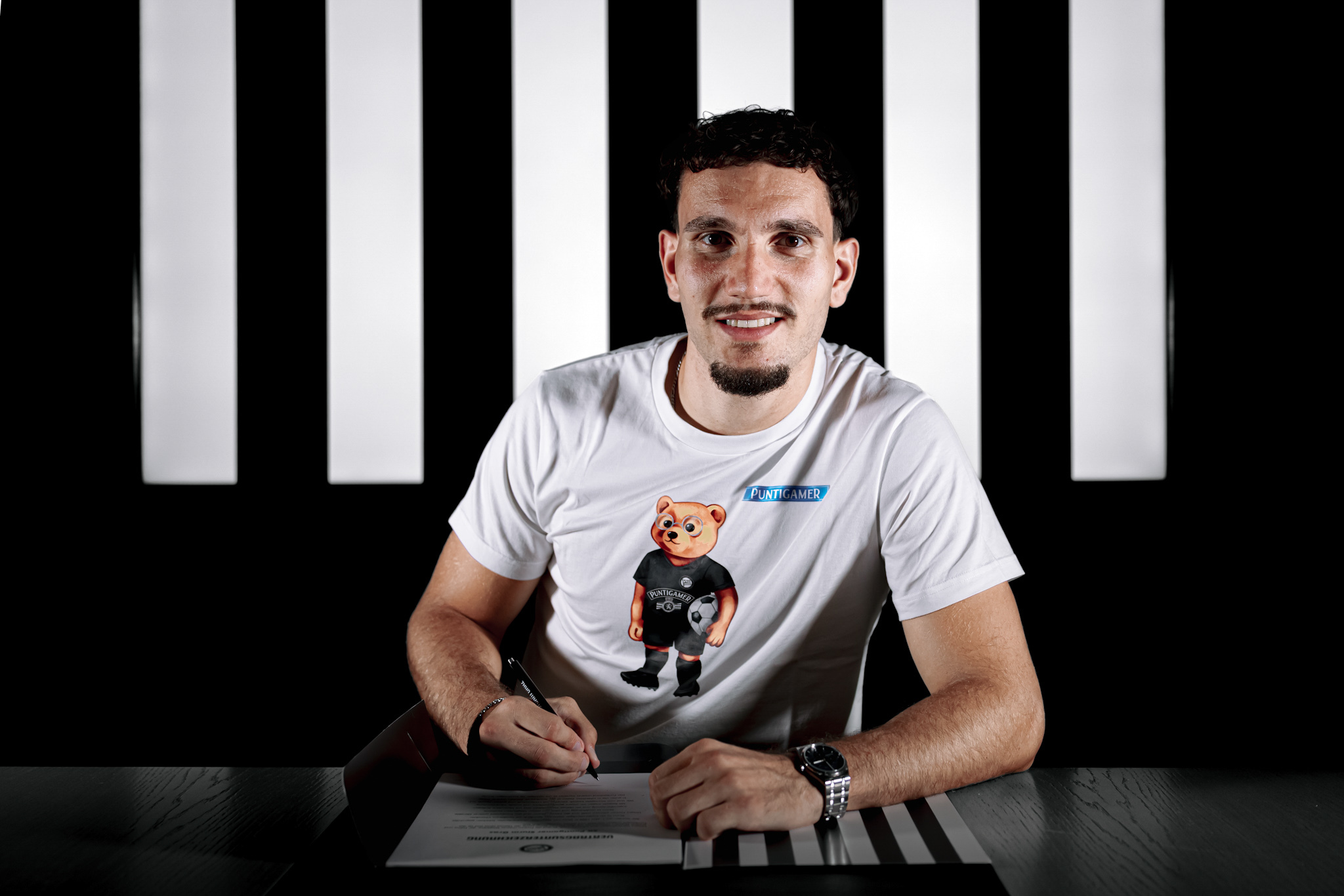 Mann trägt weißes Shirt mit Bär darauf und unterschreibt ein Blatt Papier.