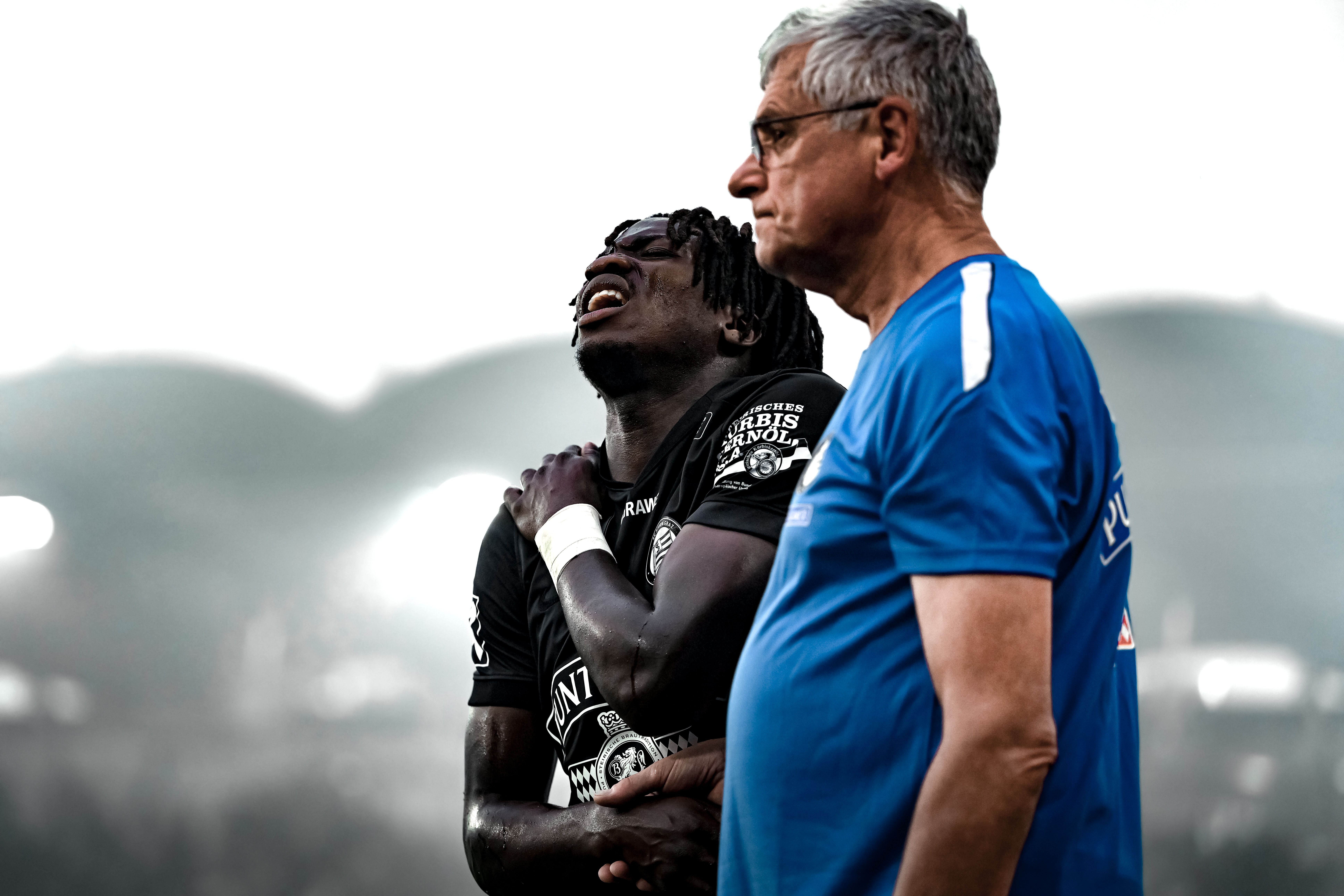 Mann in Fußballtrikot greift sich mit schmerzverzerrtem Blick an die Schulter, daneben steht ein anderer Mann.