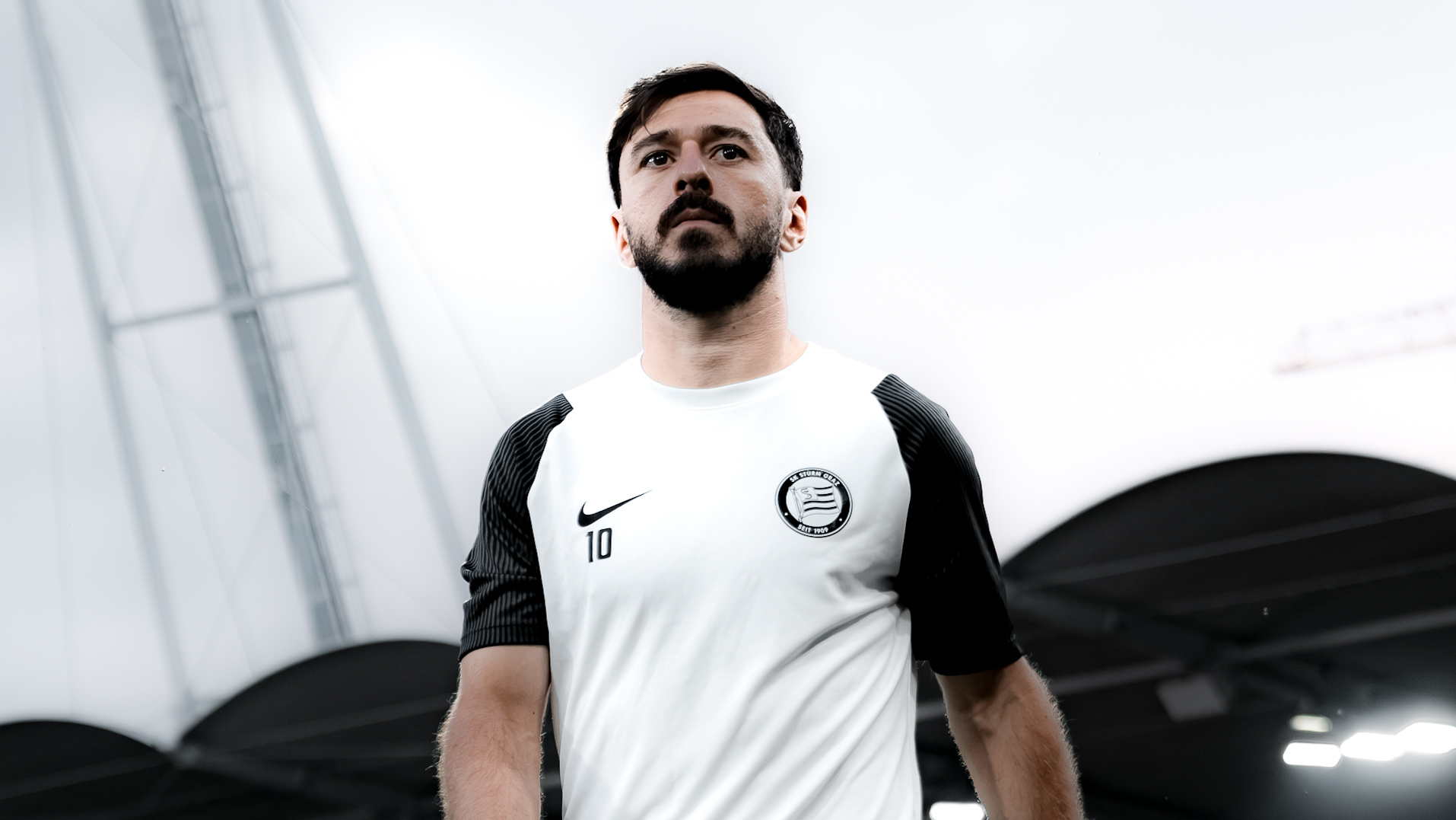 Mann in weißem Trainingsshirt blickt ernst an der Kamera vorbei.