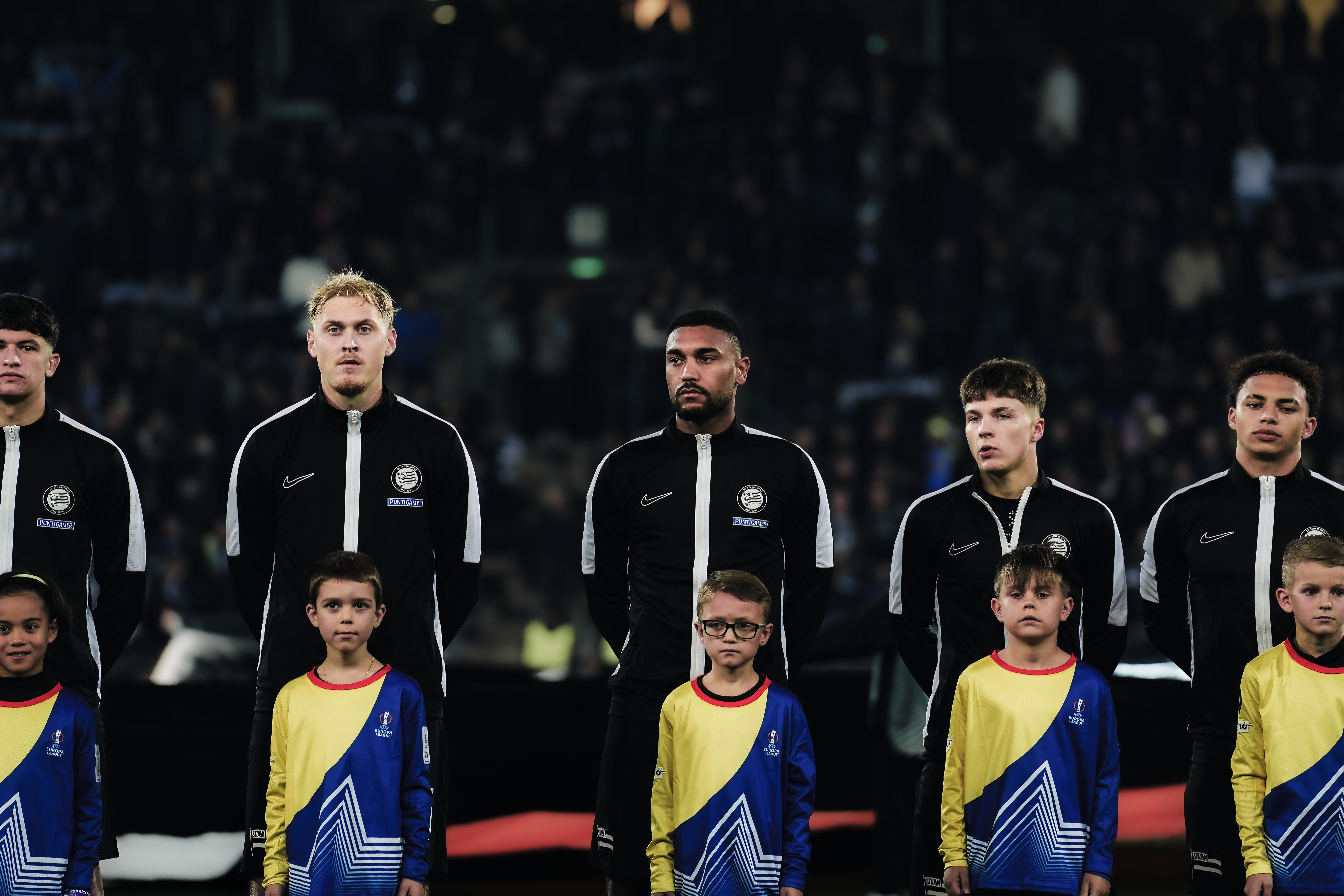 Fünf Männer mit schwarzen Westen stehen und schauen in eine Richtung und fünf Einlaufkinder stehen davor