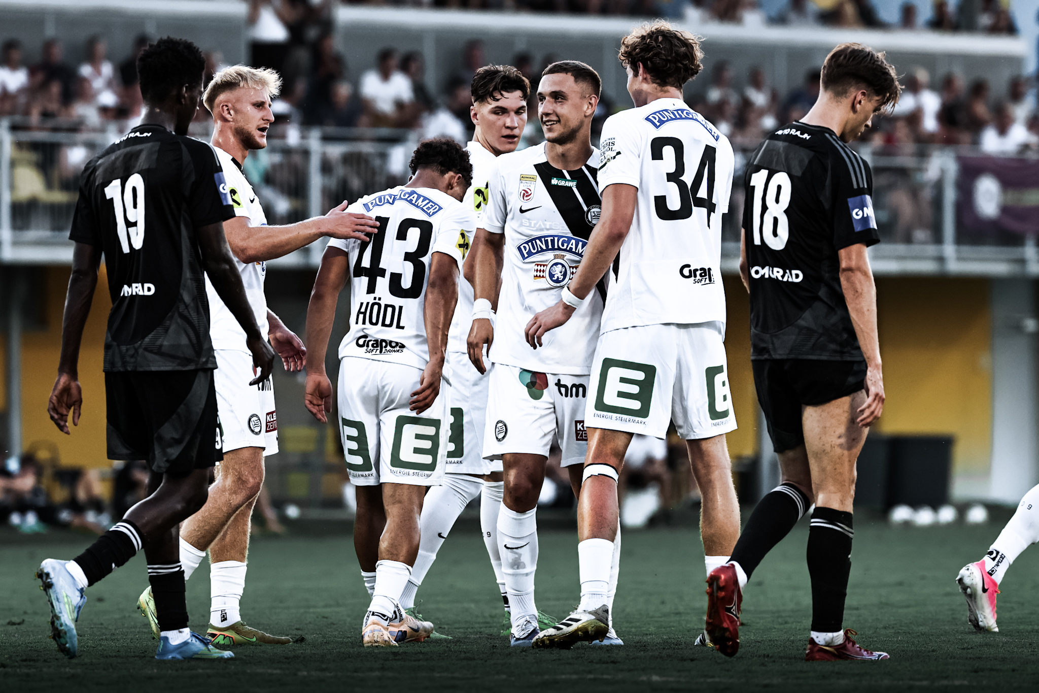 Fünf Spieler in weiß freuen sich und klatschen ab, zwei in schwarz haben hängende Köpfe. Im Hintergrund sitzen viele Menschen auf einer Tribüne.