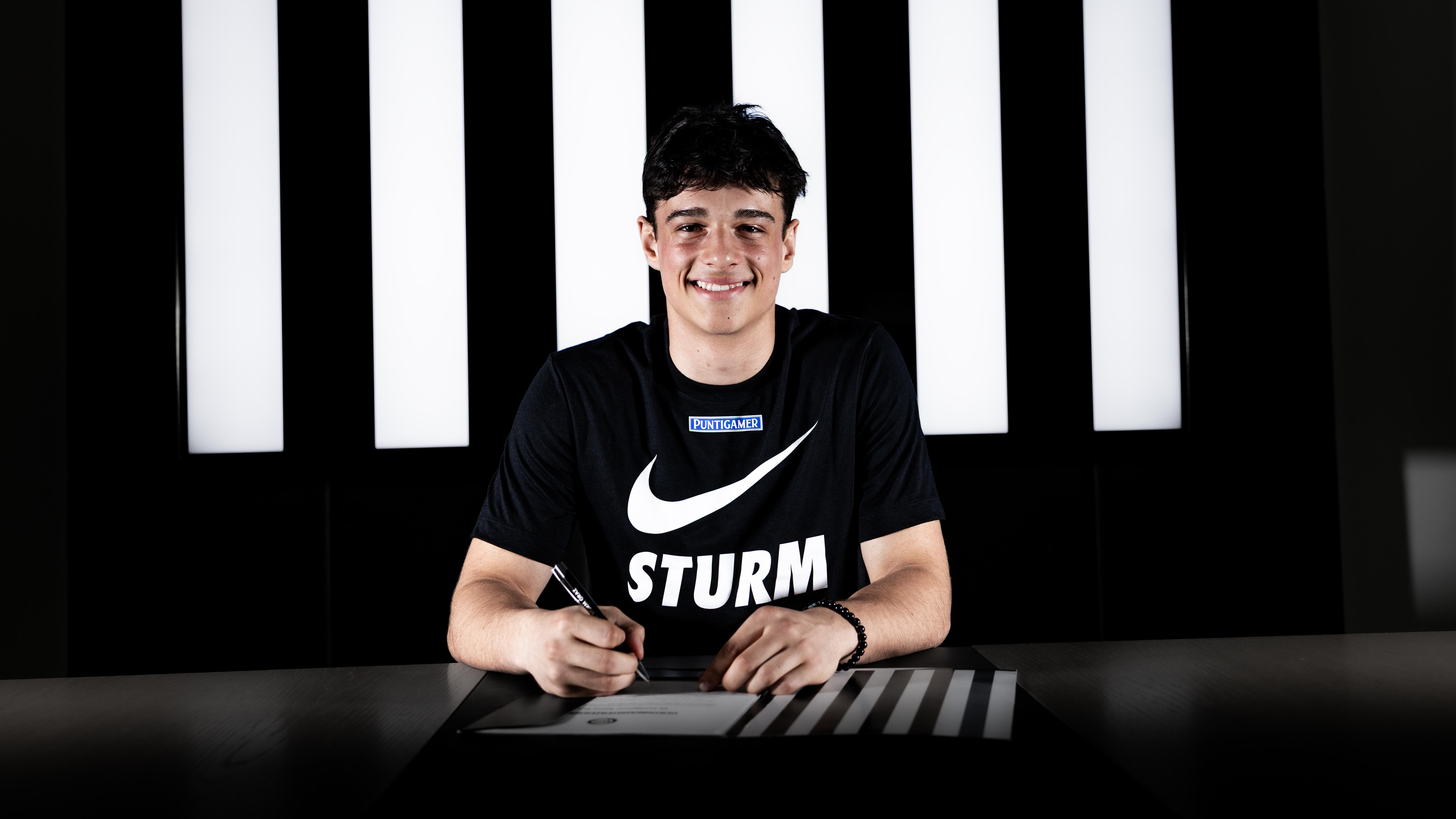 ein Mann in einem schwarzen Leiberl sitzt vor einem schwarz-weißen Bildschirm und unterschreibt mit einem Stift einen Zettel.