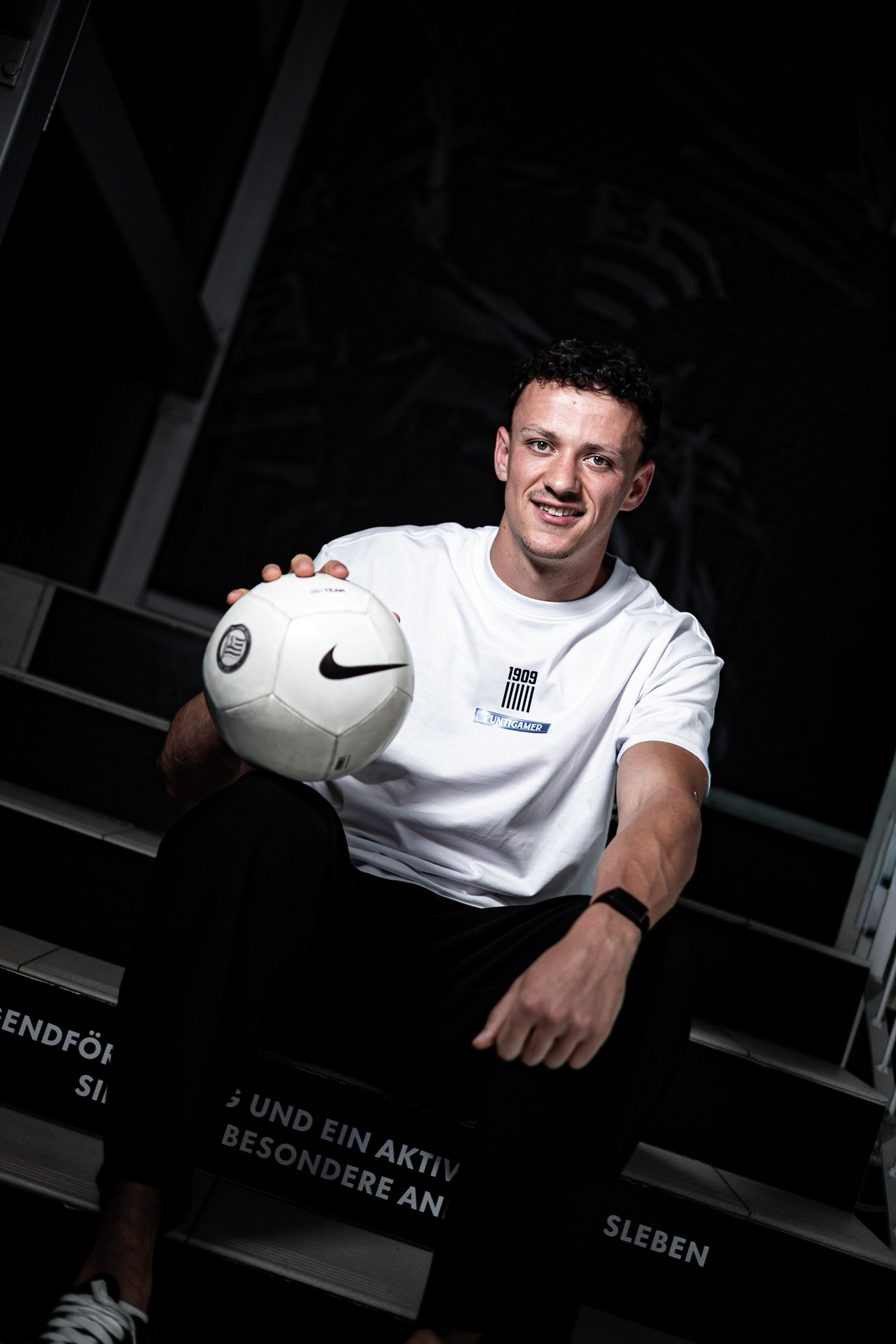Junger Mann in weißem Shirt hält weißen Ball in die Kamera.