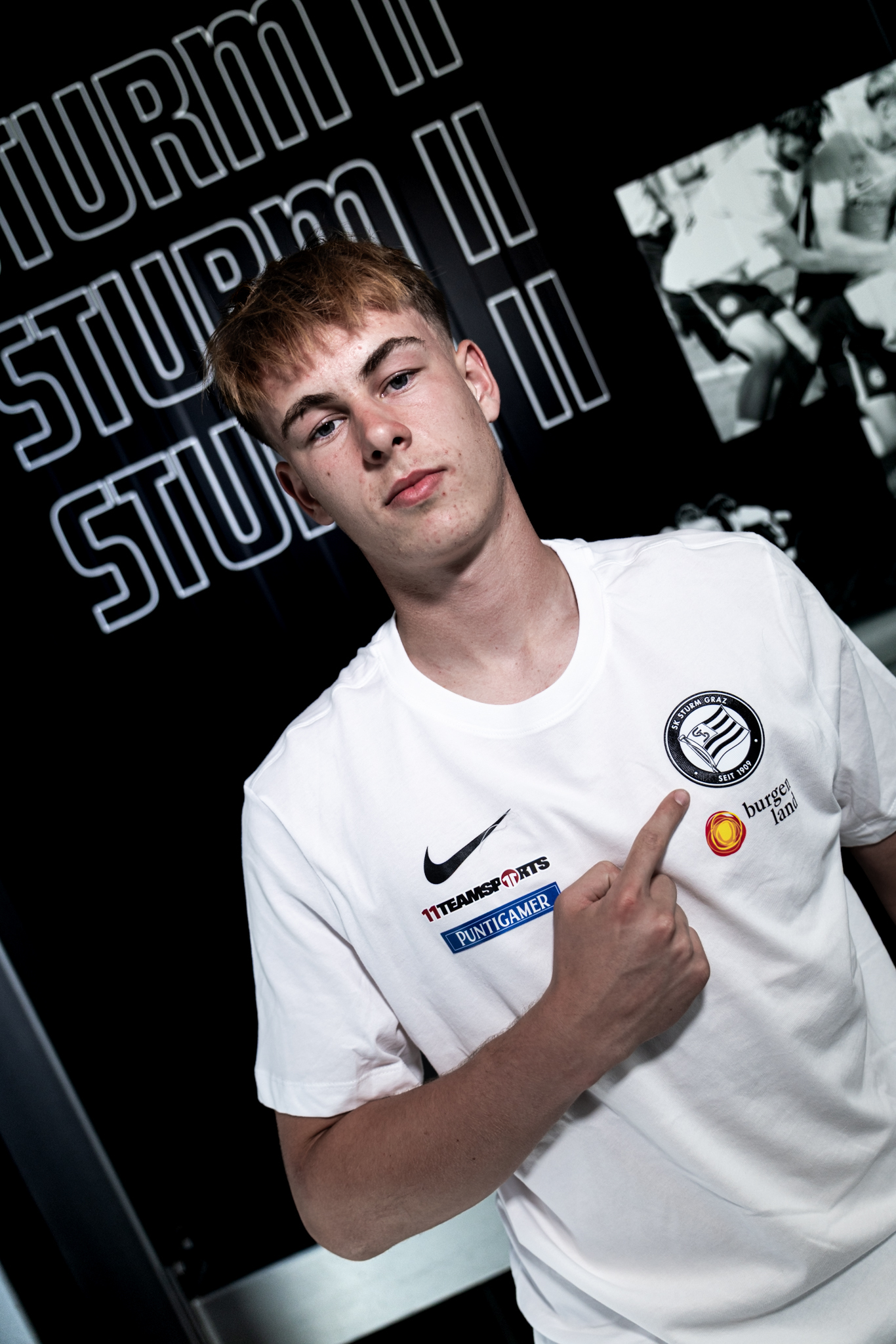 Junger Mann in weißem Shirt zeigt mit dem Finger auf ein Logo des SK Sturm auf seiner Brust.