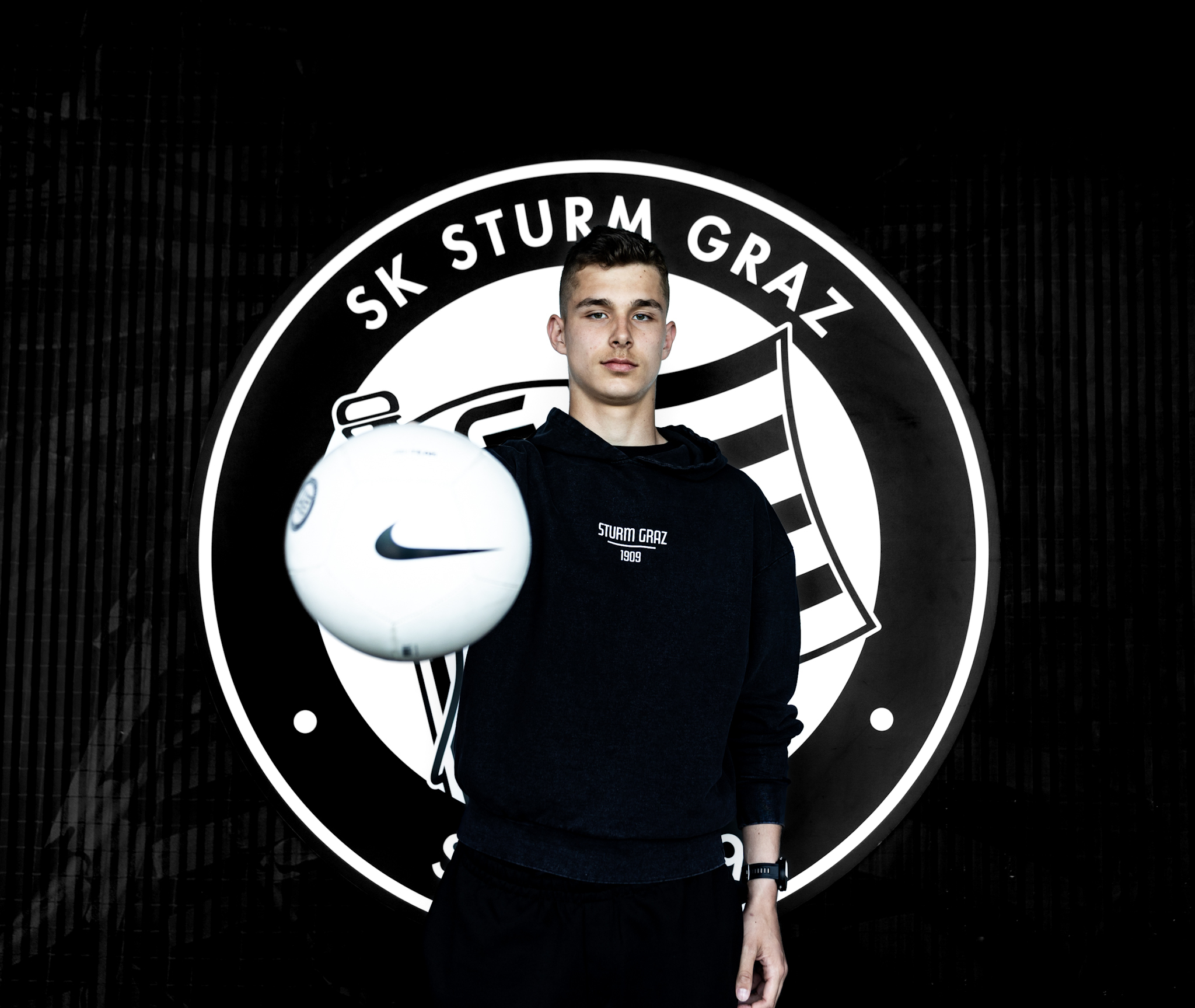 Junge in schwarzem Hoodie hält Ball in die Kamera.