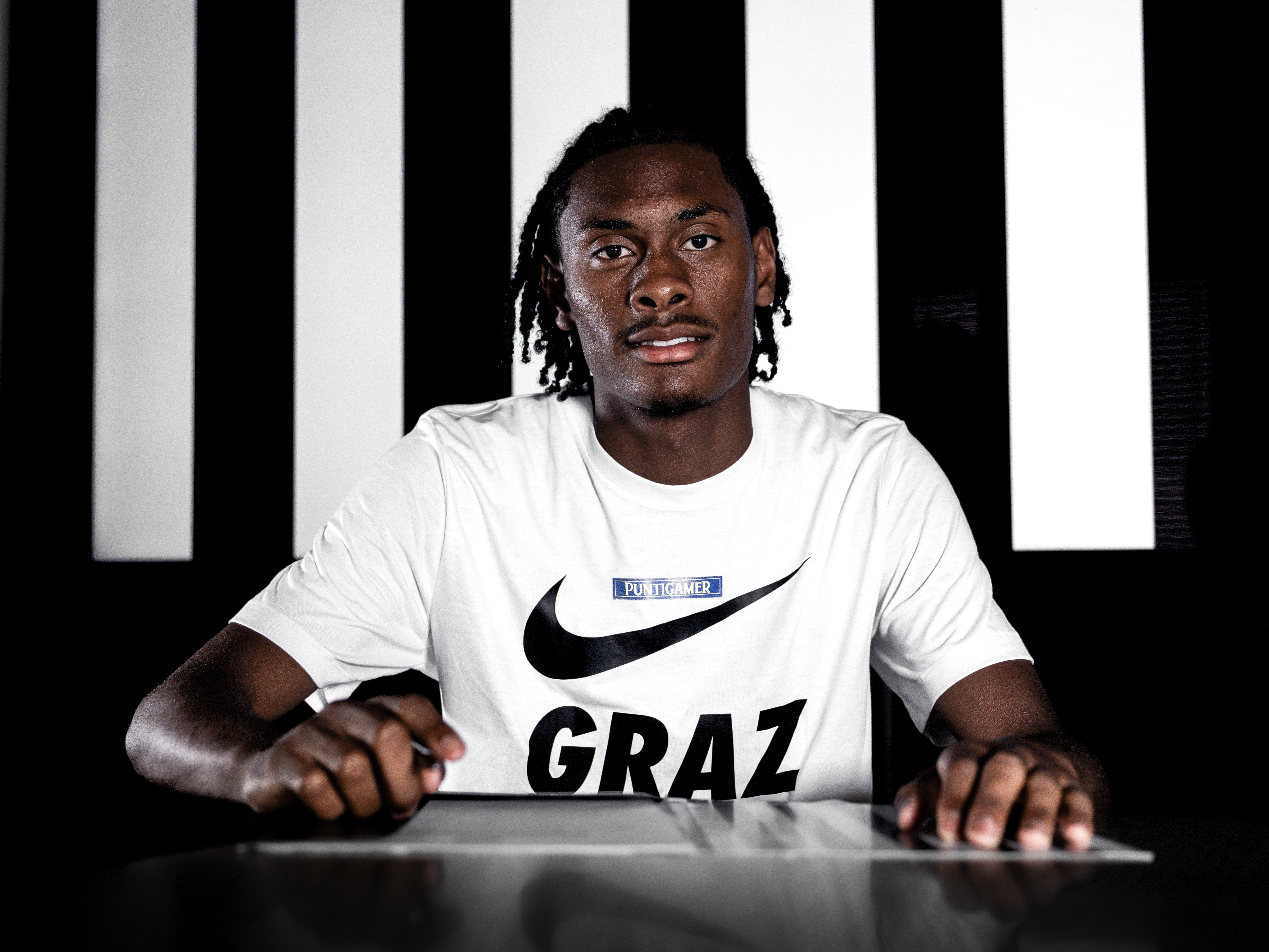 Ein Spieler in einem weißen Shirt sitzt vor einem schwarz-weißen Hintergrund und unterschreibt einen Zettel