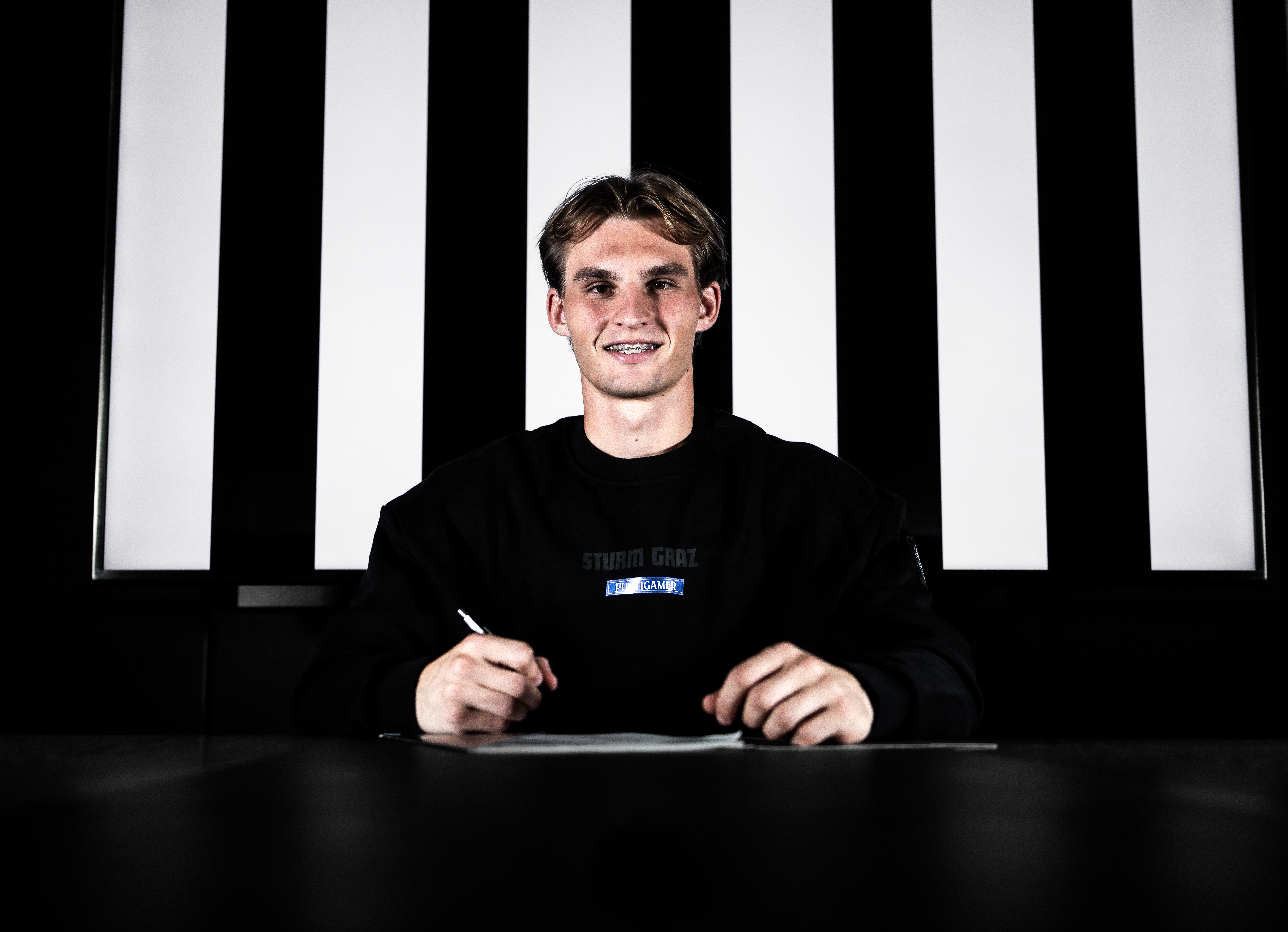 Ein Spieler in einem weißen Shirt sitzt vor einem schwarz-weißen Hintergrund und unterschreibt einen Zettel