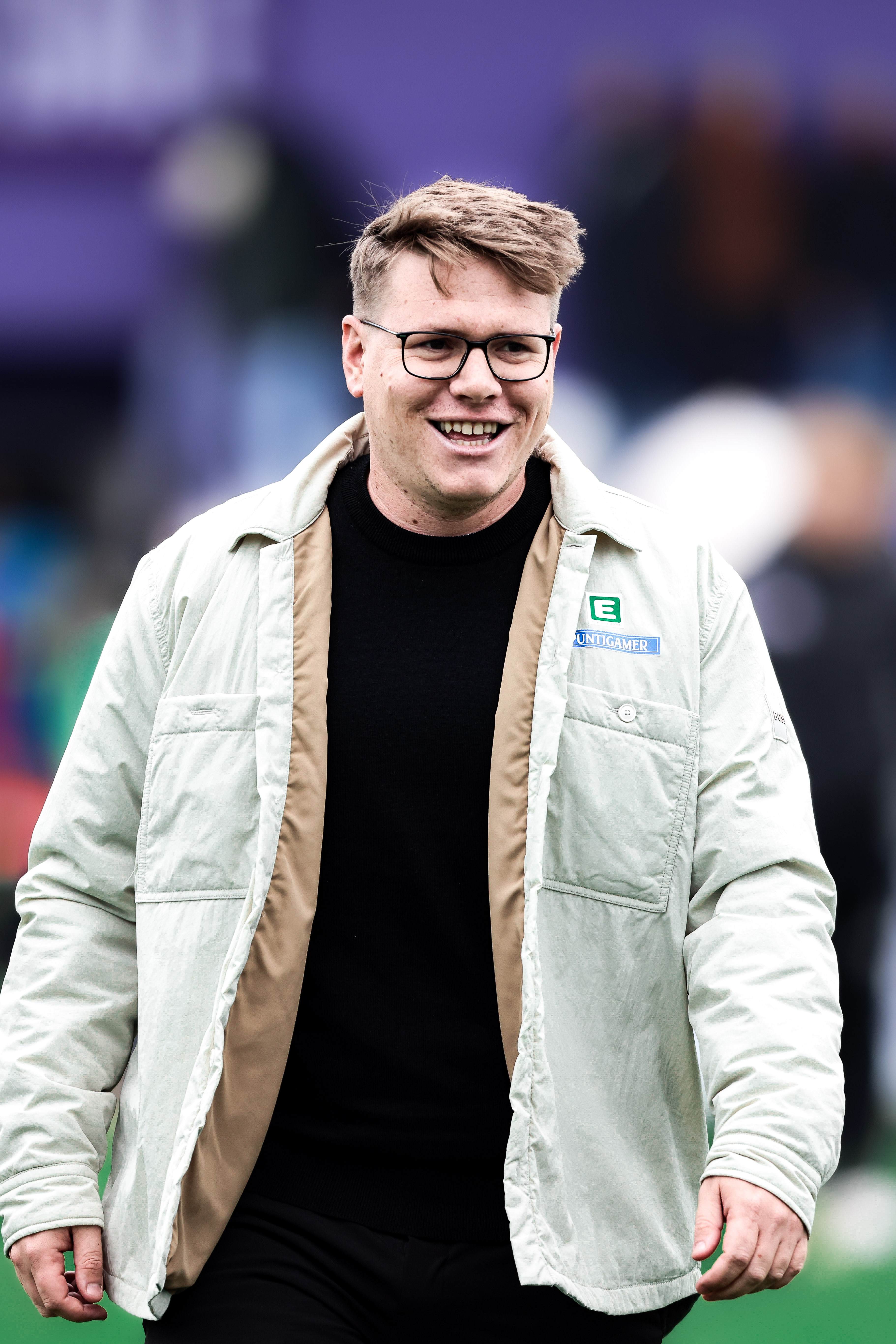 Ein Mann mit Brille und heller Jacke und dunklem Shirt lacht