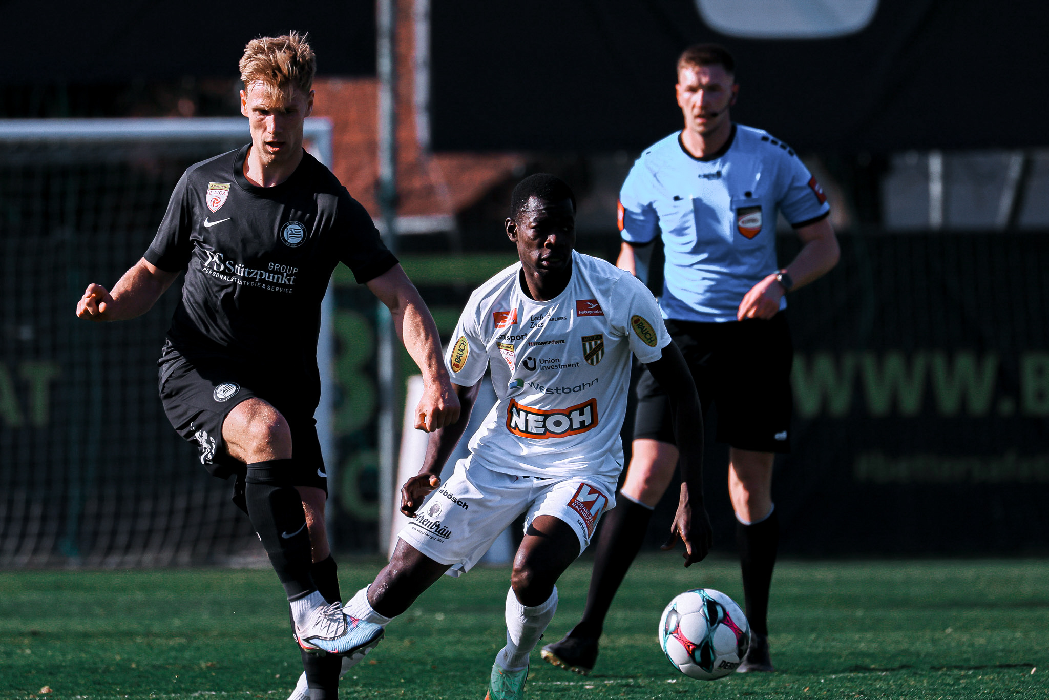 Zwei Männer spielen Fußball. Schiedsrichter schaut zu.