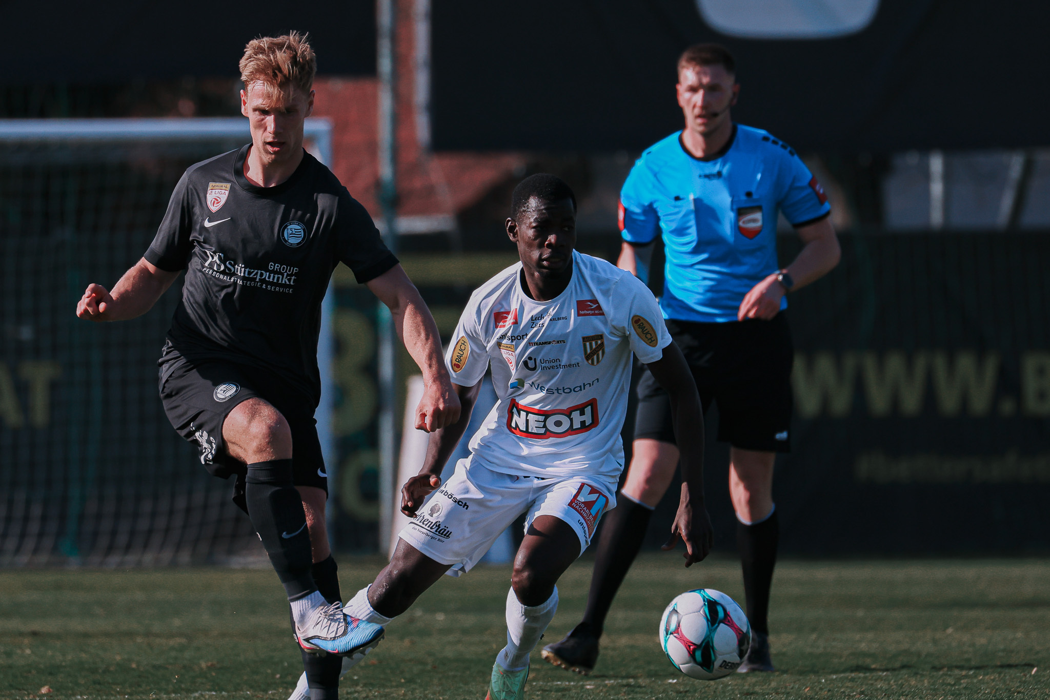 Zwei Männer spielen Fußball. Schiedsrichter schaut zu.