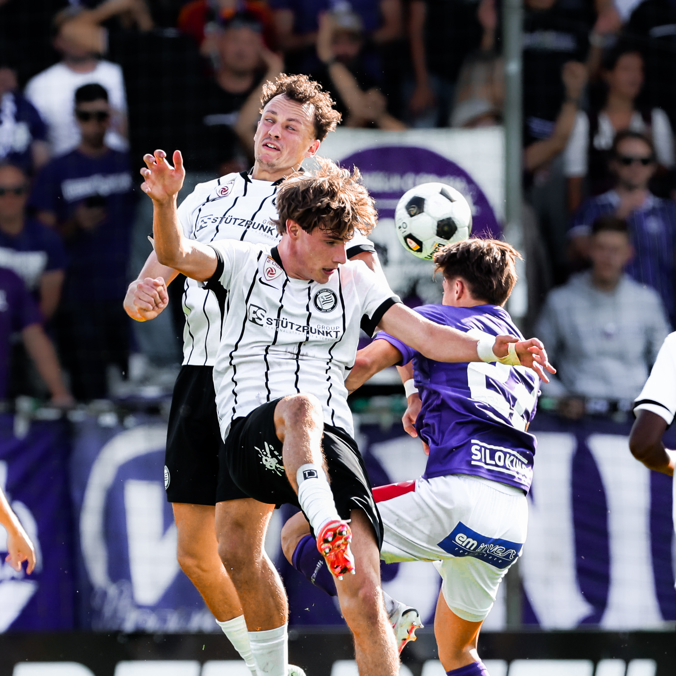 Zwei Spieler in einem weißen Trikot kämpfen mit einem Spieler in violett um einen weißen Ball.