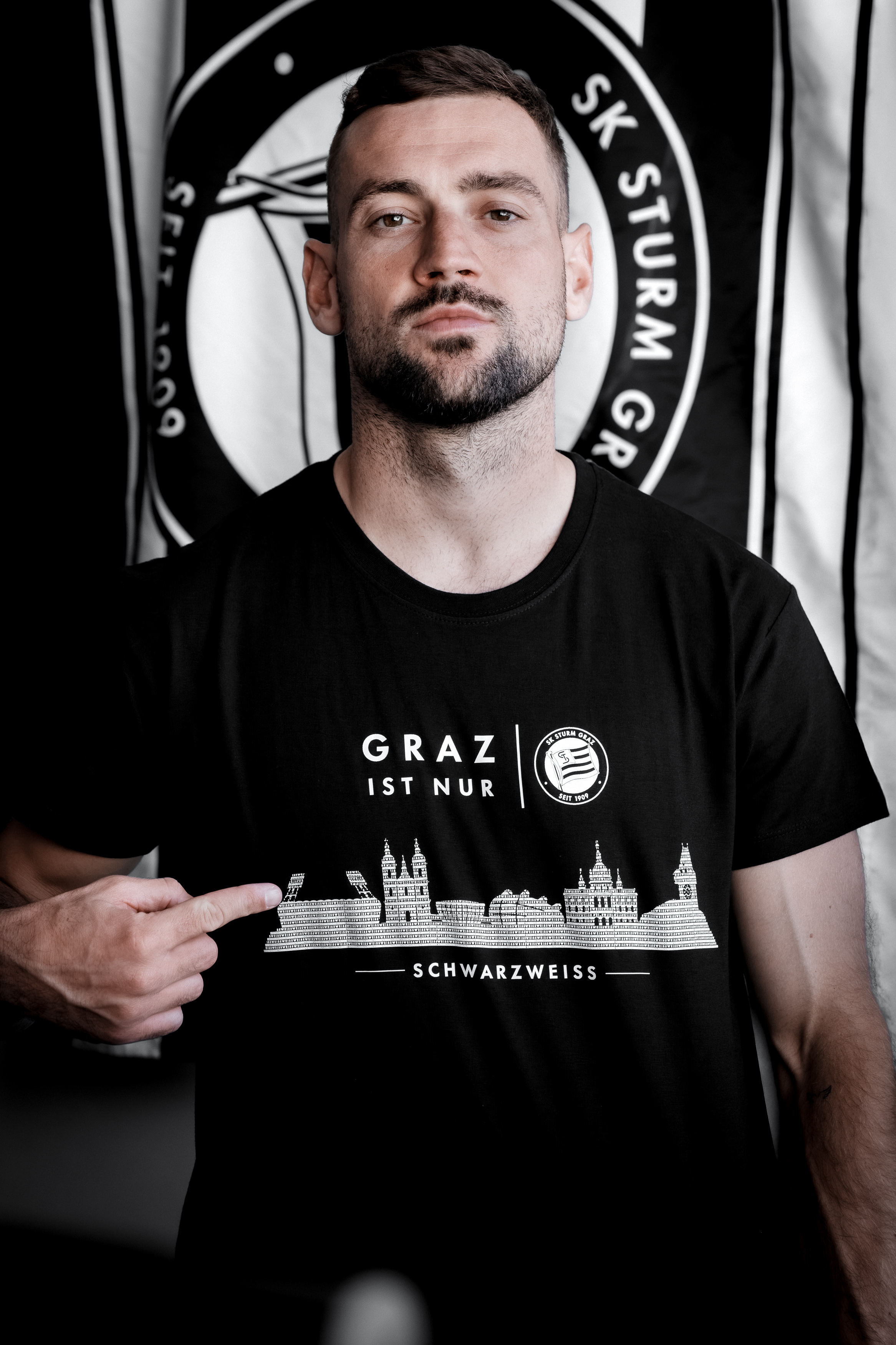 Eine Person steht mit schwarzem Shirt vor einer Fahne mit Sturm-Logo