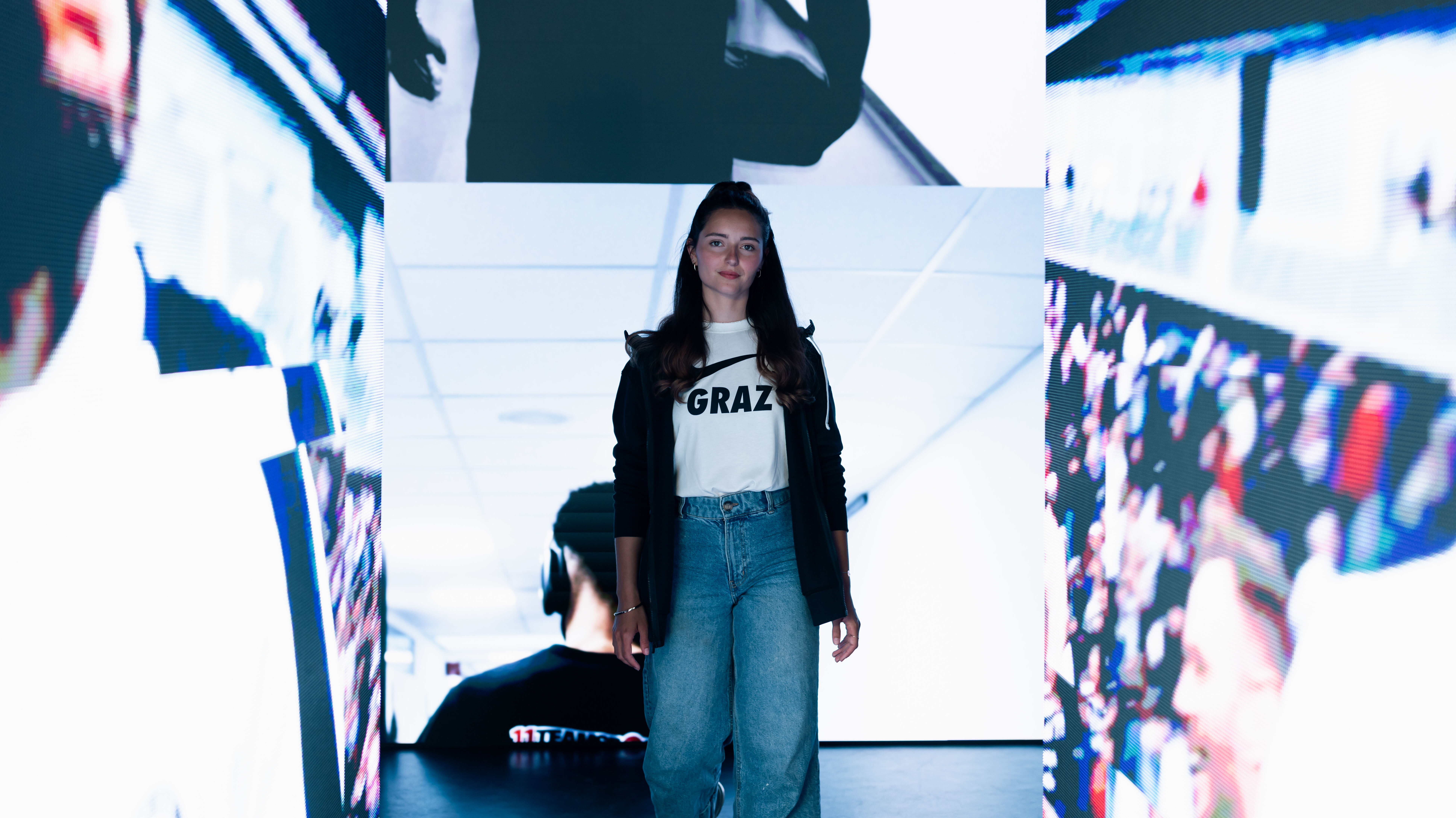 Eine junge Frau im weißen Shirt steht vor einem Screen